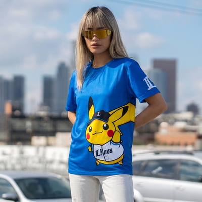 A woman wearing a bright blue short-sleeve t-shirt with a large Pikachu graphic on the front, standing outdoors with a blurred city skyline and parked cars in the background.